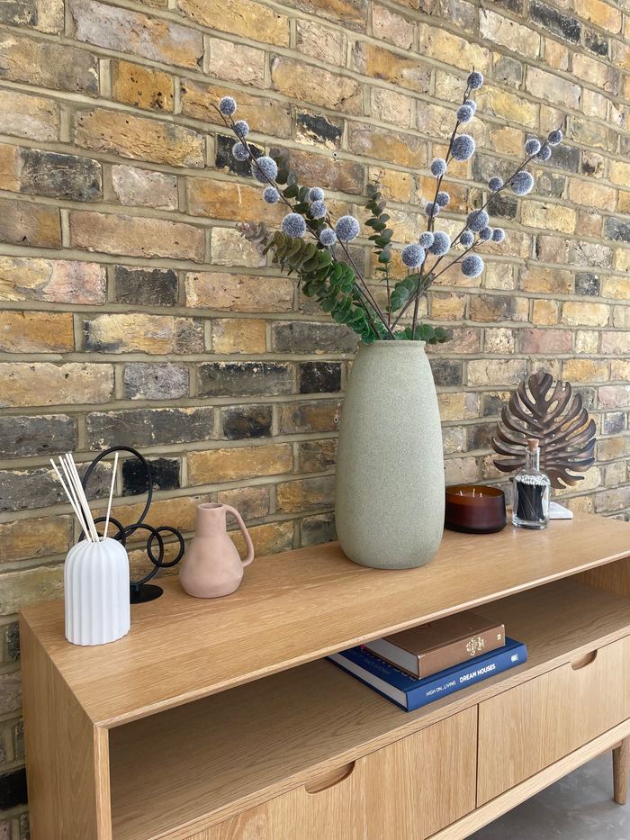 Console styling detail with a ceramic vase, dried thistle and monstera wall art against exposed brick